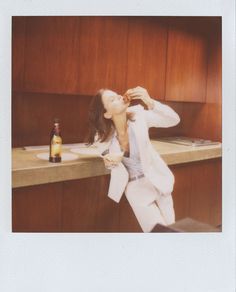 a woman sitting at a counter drinking from a bottle while wearing a white blazer