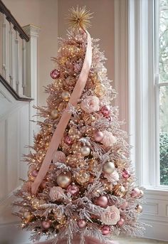 a pink christmas tree with gold ornaments and a ribbon on it in front of a window