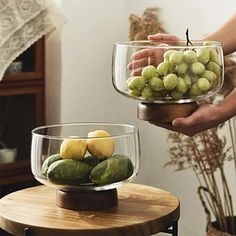 two people holding bowls filled with fruit on top of a wooden table next to each other