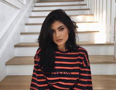 a woman with long black hair sitting in front of some stairs and looking at the camera