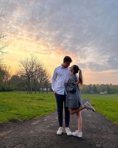 a man and woman walking down a dirt road in front of a grassy field at sunset