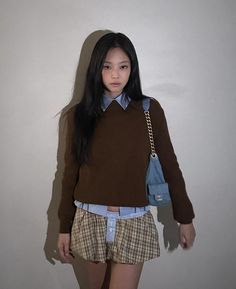 a young woman standing in front of a white wall wearing a brown sweater and plaid shorts