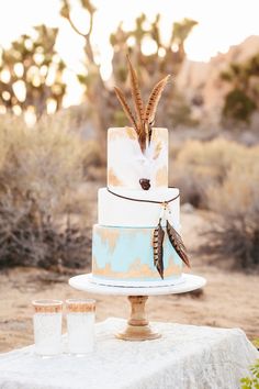 a three tiered cake with feathers on top sits on a table in the desert