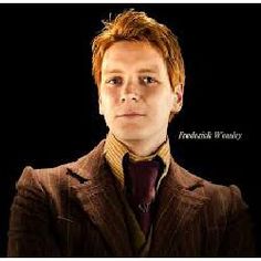 a man with red hair wearing a brown suit and tie, standing in front of a black background