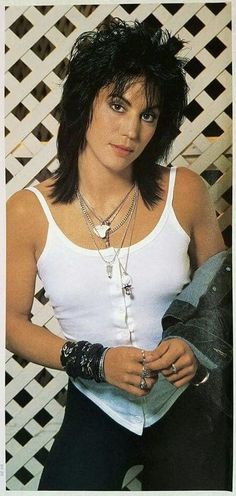 a woman with black hair wearing a white tank top and bracelets standing in front of a fence