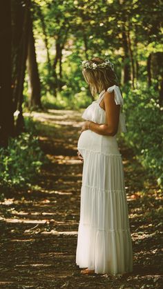 a pregnant woman in a white dress is standing on a dirt path with trees behind her