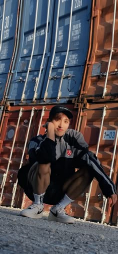 a young boy crouches in front of stacks of shipping containers, with his foot on the ground