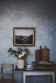 a painting hangs on the wall next to a table with a vase filled with flowers