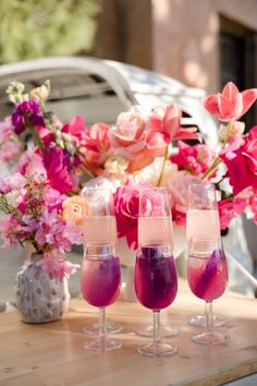 three glasses filled with liquid sitting on top of a wooden table next to pink flowers