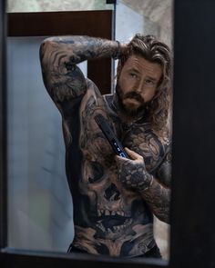 a man with long hair and tattoos is looking at his cell phone in the mirror