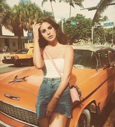 a woman standing next to an orange car on the street with palm trees in the background