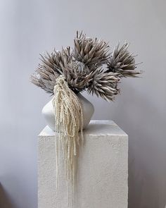 a white vase filled with dried plants on top of a block of concrete in front of a gray wall