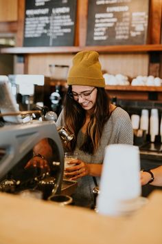 a woman in a beanie smiles as she pours her coffee at the counter