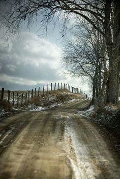 a dirt road with snow on the ground and bare trees along side it, in front of a fence