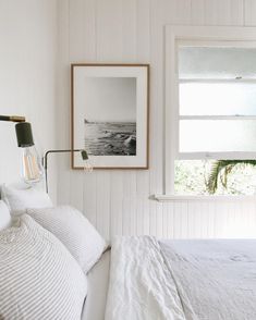 a bed with white sheets and pillows in a bedroom next to a window, framed photograph on the wall