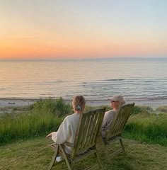 two people sitting on lawn chairs watching the sunset at the beach with their backs to each other