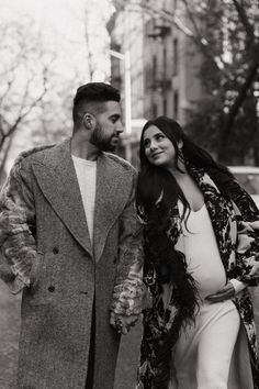 a pregnant woman walking next to a man in a long coat and feathered shawl