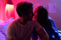 a man and woman sitting on a bed in a room with red light coming from the ceiling