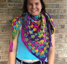 a woman standing in front of a brick wall wearing a multicolored crocheted scarf