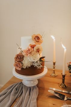 a three tiered cake with flowers on top sitting on a table next to two candles