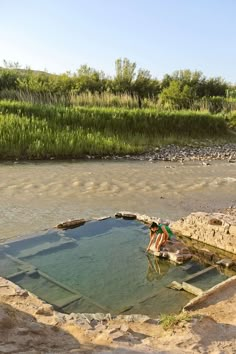 a woman is standing in the water next to some rocks and grass, near a body of water