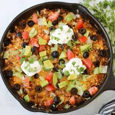 a skillet filled with mexican food and garnished with avocado, tomatoes, black olives, sour cream and sour cream