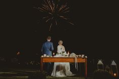 a bride and groom standing at a table with fireworks in the background