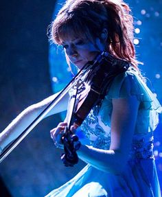 a woman holding a violin on stage in front of a microphone and wearing a blue dress