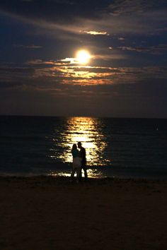 two people are standing on the beach at night with the moon in the sky above them