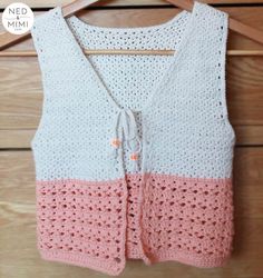 a pink and white crocheted vest hanging on a wooden hanger next to a wood wall