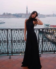 a woman in a long black dress standing on a balcony overlooking the water and boats