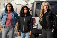 three young women standing next to each other in front of a car with graffiti on the wall behind them