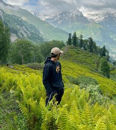 a man standing on top of a lush green hillside covered in trees and bushes with mountains in the background