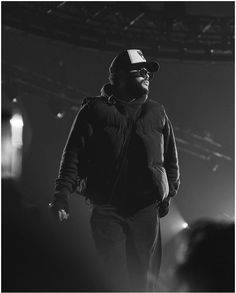 a black and white photo of a man on stage wearing a baseball cap, jacket and sunglasses