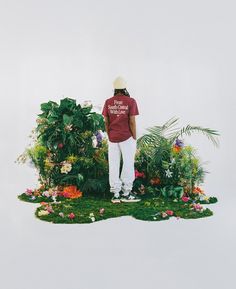 a man standing on top of a lush green field next to flowers and plants in front of a white wall