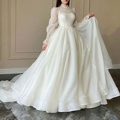 a woman wearing a white wedding dress with long sleeves and sheer neckline, standing in front of a wall