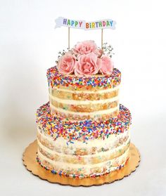 a three layer cake with sprinkles and flowers on top is decorated with a happy birthday sign