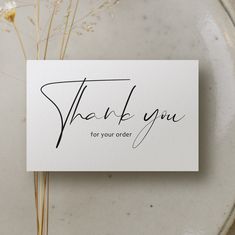 a thank you card sitting on top of a white plate next to some dried flowers