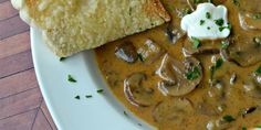 a white plate topped with meat and mushrooms covered in gravy next to a piece of bread