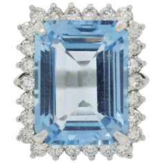 A large and luxurious blue topaz ring! The sky-blue colored gem weighs 25.69 carats and has a precise emerald-cut and rectangular shape that is free of any eye-visible inclusions. It is accented by 1.99 carats of round brilliant-cut diamonds which halo the Swiss blue topaz and add brilliance to the piece. Hand-fabricated in platinum and ready to make a statement! (Swiss topaz is the trade name for bright blue topaz, while London blue is used for the darker blue stones) Ring Size 6.25 Cocktail Ring Diamond, Blue Stone Ring, Colored Gems, Swiss Blue Topaz, Blue Topaz Ring, Topaz Ring, Round Brilliant Cut Diamond, Cocktail Ring, Blue Stone