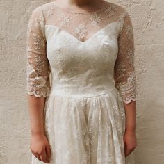 a woman standing in front of a wall wearing a white dress with sheer sleeves and lace