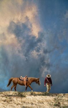 a man with a cowboy hat is leading a horse on a hill under a cloudy sky