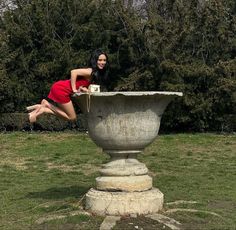 a woman in a red dress jumping into a large stone vase on the grass with trees behind her