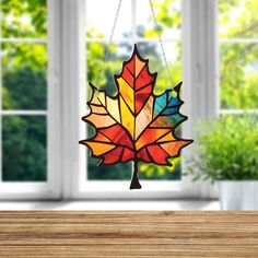 a stained glass maple leaf hanging from a chain on a wooden table in front of a window