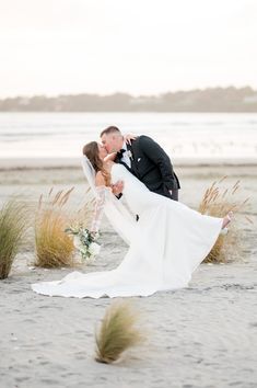 a bride and groom kissing on the beach in front of some tall grass with water behind them