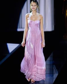 a woman walking down a runway wearing a pink dress with pleated skirt and high heels