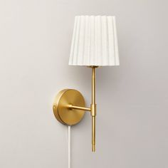 a gold wall light with a white shade on it's side and a lamp plugged into the wall