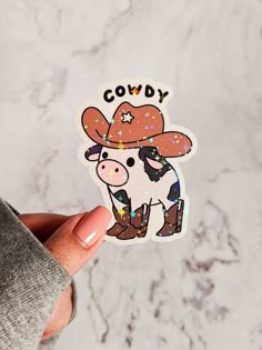 a person holding up a sticker with a cow wearing a cowboy hat on it