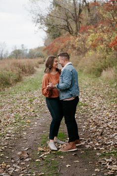 an engaged couple standing on a path surrounded by leaves and trees in the fall with their arms around each other