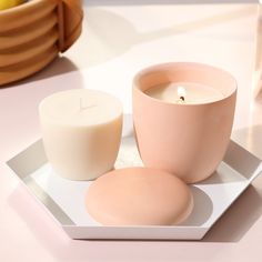 two candles on a white plate next to a pink candle holder and some bananas in the background